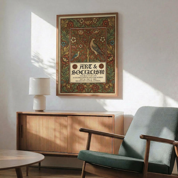 May include: Ornate Arts and Crafts style poster with intricate floral border in earthy tones of green, rust, and gold, featuring a detailed bird among flowering vines. The decorative print reads "Art & Socialism" in Gothic lettering within an elaborate botanical frame. Displayed above a mid-century modern wooden credenza with sliding doors, beside a sage green upholstered armchair with wooden frame. Warm sunlight casts geometric shadows across white walls.