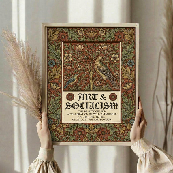 May include: Hands holding a framed vintage-style poster featuring ornate floral border design with birds, in rich burgundy, gold and green tones. Gothic-style text reads "ART & SOCIALISM" with decorative flowers and detailed venue information below. Light wooden frame against soft cream curtains with dried pampas grass visible to the left side.