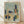 May include: Framed abstract architectural artwork in light wood frame leaning against white wall on wooden floor. Watercolor-style painting features geometric buildings, trees, and urban landscape in muted blues, oranges, greens, and beiges. Modernist cubist composition with overlapping shapes and soft brushwork creates dreamlike cityscape scene.