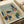 Abstract modernist artwork featuring geometric buildings and architectural forms in muted blues, oranges, greens, and beige tones. The composition shows angular structures, including a tall dark blue tower, houses with linear details, and a stylized tree, all arranged in a cubist style. The print appears aged with subtle staining and is displayed in an open art book or portfolio with cream-colored pages on a white surface.
