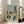 May include: Framed abstract architectural artwork leaning against white wall next to beige sofa. The print features geometric buildings, trees, and structures in muted blue, green, orange, and cream tones with cubist-style composition. Light wood frame, positioned on hardwood floor in bright room with large windows.