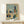 May include: Abstract modernist architectural artwork in wooden frame displaying geometric buildings, trees, and cityscape in muted blues, oranges, and greens. Watercolor-style painting with cubist influence showing angular structures and organic forms. Frame sits on light wood surface beside small white wrapped gift with evergreen sprig against cream wall.
