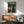 May include: Framed anime-style poster featuring a dark-haired figure in brown coat standing on balcony overlooking neon-lit cyberpunk cityscape with Japanese text at top, displayed above wooden sideboard with white ceramic lamp in modern living room with mint green upholstered chair and small round coffee table, natural sunlight casting shadows on white wall