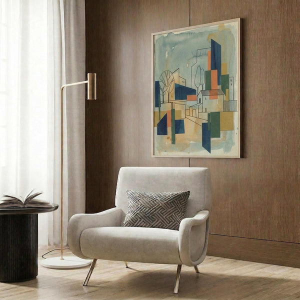 May include: Framed abstract architectural artwork featuring geometric buildings, trees, and structures in blue, orange, green, and beige watercolor tones, displayed on wood-paneled wall above cream upholstered armchair with patterned pillow, beside brass floor lamp and dark round side table with open book.