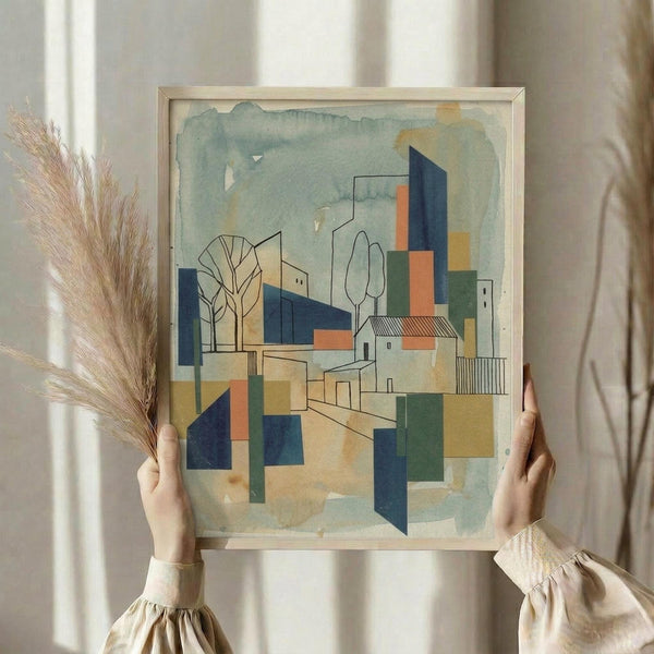 May include: Person holding framed abstract architectural artwork featuring geometric buildings, trees, and overlapping shapes in blues, greens, oranges and yellows with watercolor-style background. Light wood frame against white curtained window with dried wheat stems visible. Artwork shows cubist-style cityscape with angular buildings and simplified tree forms.