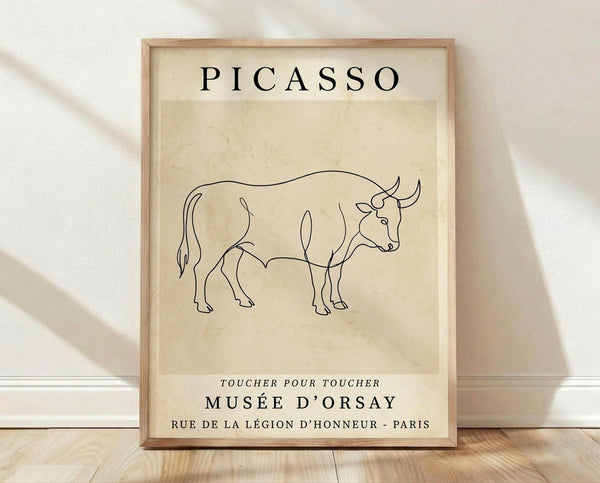 May include: Framed poster with cream textured background featuring minimalist black line drawing of a bull in Picasso's continuous line style. Text reads "PICASSO" at top and "MUSÉE D'ORSAY" with French museum details below. Light wood frame sits on wooden floor against white textured wall with soft natural lighting creating diagonal shadows.