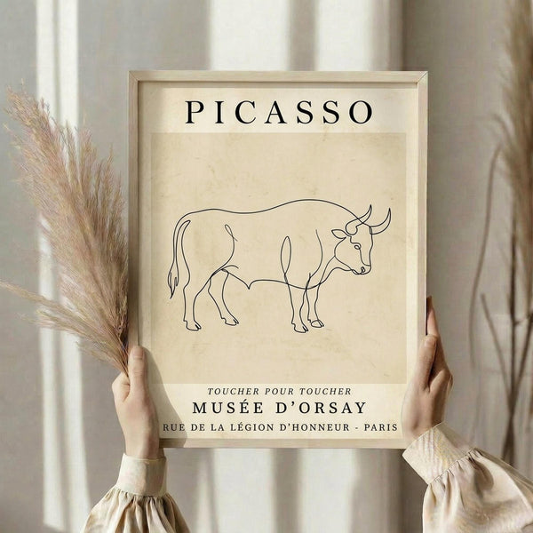 May include: Person in cream ruffled sleeves holding framed poster and dried pampas grass against white curtained window. Beige-framed print features minimalist black line drawing of bull on cream background with "PICASSO" at top and French museum text below. Soft natural lighting creates warm, neutral aesthetic.