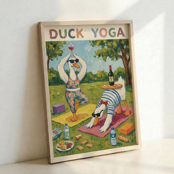 A framed illustration showing two ducks practicing yoga in a park setting. One duck in floral yoga attire holds a wine glass while in tree pose, the other wears a striped shirt doing downward dog on a pink mat. The colorful scene includes scattered snacks, wine bottles, yoga blocks, and water bottles among green trees. The words "DUCK YOGA" appear at the top in multicolored letters. Light wooden frame against white wall.