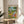 May include: Framed poster print displayed on light wood round side table next to beige upholstered chair. Artwork shows two cartoon ducks doing yoga outdoors - one duck in colorful leggings holding wine glass in tree pose, another in striped shirt doing downward dog on pink yoga mat. Scene includes wine bottle, baguette, cheese, yoga blocks, and green park setting. Natural lighting creates soft shadows across white wall background.