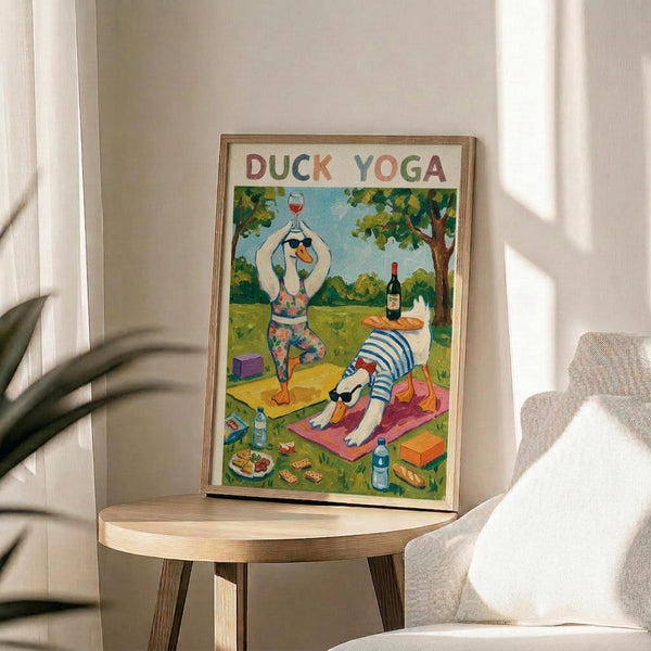 May include: Framed poster print displayed on light wood round side table next to beige upholstered chair. Artwork shows two cartoon ducks doing yoga outdoors - one duck in colorful leggings holding wine glass in tree pose, another in striped shirt doing downward dog on pink yoga mat. Scene includes wine bottle, baguette, cheese, yoga blocks, and green park setting. Natural lighting creates soft shadows across white wall background.