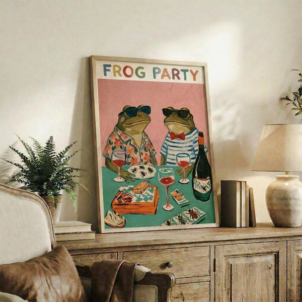 May include: Framed poster showing two frogs wearing sunglasses and casual shirts sitting at a turquoise table with wine glasses, cheese, and snacks under colorful "FROG PARTY" text on coral pink background. Wood-framed print displayed on rustic wooden sideboard alongside books, ceramic vase, and fern plant in cozy living room setting with cream sofa and neutral walls.