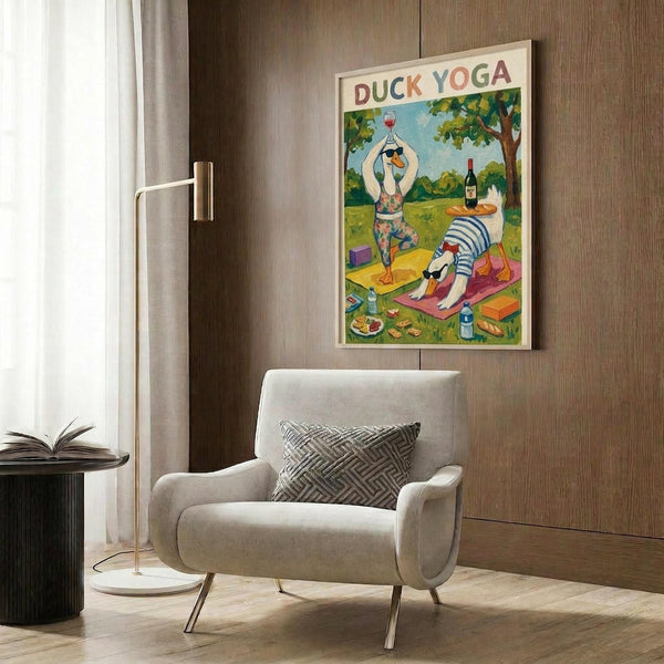May include: Framed poster showing two cartoon ducks doing yoga poses outdoors, with wine glasses and French bread nearby. The colorful illustrated print hangs on wood-paneled wall above a cream-colored modern armchair with geometric pillow. Room features gold floor lamp and dark side table with open book.