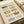 May include: Watercolor illustration print displayed in cream matted frame showing collection of vintage casserole dishes and baking pans in soft pastels. Features floral-patterned cookware in green, pink, yellow, orange, and blue tones with delicate botanical motifs. Print sits on neutral surface beside rolled paper, showcasing nostalgic mid-century kitchen aesthetic with gentle brushwork textures.