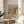 May include: Framed poster displaying two anthropomorphic dogs having tea - a poodle in floral dress holding teacup and bulldog in vest with bow tie. Table set with tiered serving plates, sandwiches, scones, floral teapot and china cups. Warm yellow and green color palette. Wood frame rests on round wooden side table next to beige upholstered chair, with plant leaves visible and soft window lighting.