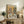 May include: Framed vintage-style poster showing two anthropomorphic dogs having tea - a poodle in floral dress and bulldog in green vest with bow tie, sitting at table with fine china, teacups, scones and sandwiches. Poster displayed on rustic wooden sideboard with books, ceramic vase, and fern plant in cozy living room setting with beige armchair visible.