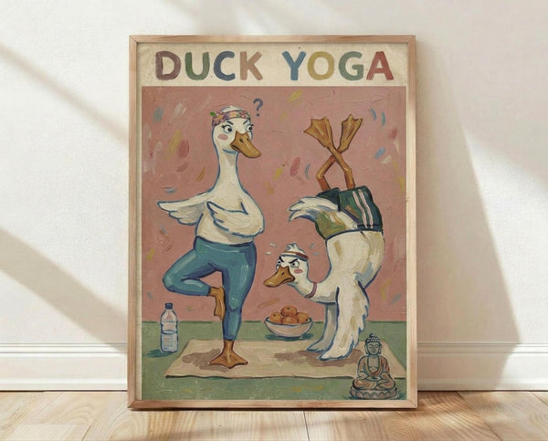 May include: Framed poster print leaning against white wall on light wooden floor. Artwork shows two cartoon ducks doing yoga poses on a mat - one in tree pose wearing blue leggings and headband, another in crossed-legs position. Pink background with colorful brushstrokes, water bottle and bowl nearby, small Buddha statue in corner. Natural wood frame, soft lighting creates shadows on textured white wall.