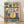 May include: Framed colorful folk art style kitchen illustration showing three shelves with potted plants, hanging utensils, teapots, mugs, and decorative bowls. Features bright blues, yellows, pinks, and greens with floral and geometric patterns. Natural wood frame sits on light wooden floor against white wall with soft shadows.