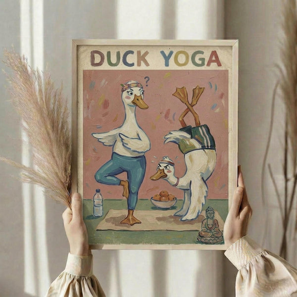 May include: Hands holding a framed poster showing cartoon ducks doing yoga poses on a yoga mat. One duck in blue leggings does tree pose with a bandana and question mark, another does headstand. Scene includes water bottle, bowl of snacks, and small Buddha statue. Pink background with colorful brushstrokes. Dried wheat stems visible to left side.