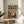 May include: Colorful folk art style kitchen illustration in wooden frame, displayed on round wooden side table next to beige upholstered chair. Artwork features decorative potted plants on shelves, hanging kitchen utensils, teapots and vessels in vibrant blues, yellows, reds and greens with whimsical patterns. Natural lighting creates soft shadows on white wall behind frame.