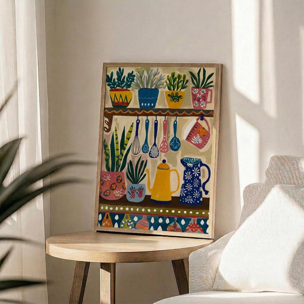 May include: Colorful folk art style kitchen illustration in wooden frame, displayed on round wooden side table next to beige upholstered chair. Artwork features decorative potted plants on shelves, hanging kitchen utensils, teapots and vessels in vibrant blues, yellows, reds and greens with whimsical patterns. Natural lighting creates soft shadows on white wall behind frame.