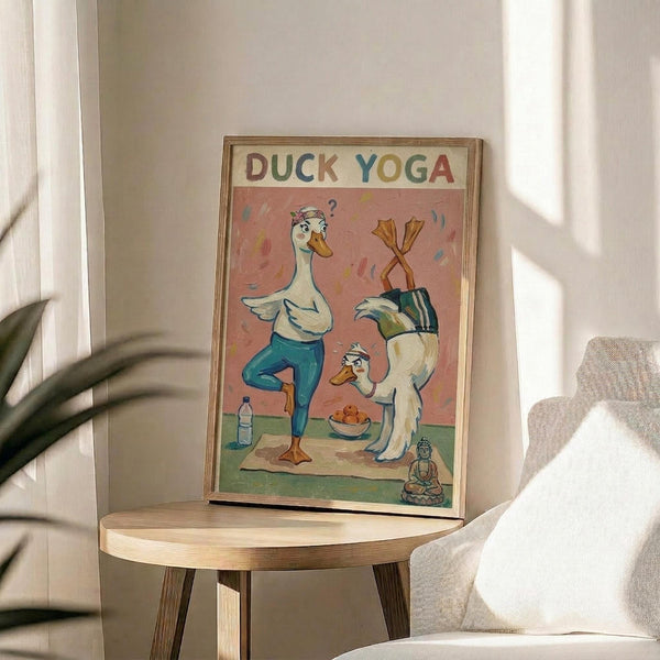 May include: Whimsical illustrated poster featuring cartoon ducks doing yoga poses on a coral pink background, with colorful "DUCK YOGA" text at top. One duck in teal pants does a standing pose while another is in headstand position. Small Buddha figure and water bottle visible on yoga mat. Framed in light wood frame, displayed on round wooden side table next to beige upholstered chair with plant shadows cast on white wall behind.
