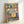 May include: Framed kitchen-themed folk art print leaning against white wall. Colorful illustration shows wooden shelves with potted plants, hanging utensils including whisk and ladles, teapots in yellow and blue with decorative patterns, and ornamental border. Artwork features warm earth tones, botanical elements, and traditional folk art styling in light wood frame.