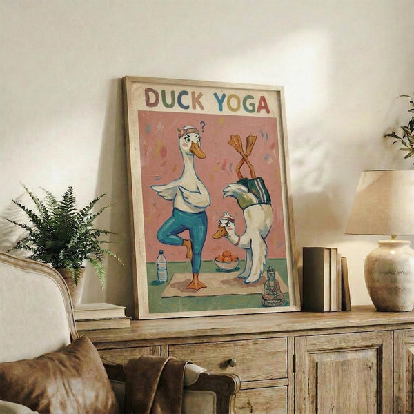 May include: Framed poster displaying cartoon ducks doing yoga poses on a coral pink background, with colorful lettering spelling "DUCK YOGA" at top. One duck in blue leggings performs tree pose while another duck sits nearby with a question mark above its head. Small Buddha statue and water bottle visible in scene. Frame sits on rustic wooden sideboard alongside potted fern, books, ceramic vases, and brown leather chair in cozy living room setting with white walls.