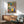 May include: Colorful folk art style kitchen poster displayed in modern living room with mid-century furniture. Artwork shows decorative shelf with potted plants, hanging utensils, coffee pots and kitchenware in vibrant blues, yellows, pinks and greens. Framed print hangs above wooden sideboard next to table lamp, with gray upholstered chair visible. Warm natural lighting creates shadows on white wall.