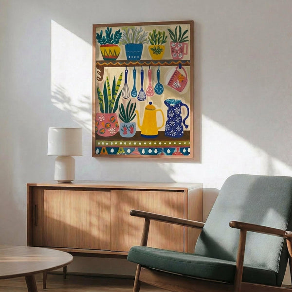 May include: Colorful folk art style kitchen poster displayed in modern living room with mid-century furniture. Artwork shows decorative shelf with potted plants, hanging utensils, coffee pots and kitchenware in vibrant blues, yellows, pinks and greens. Framed print hangs above wooden sideboard next to table lamp, with gray upholstered chair visible. Warm natural lighting creates shadows on white wall.
