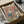May include: Vintage-style poster artwork featuring two cartoon ducks doing yoga poses, titled "Duck Yoga" in colorful letters. One duck wears teal leggings while stretching, the other duck sits nearby with a bowl of snacks. The illustration has warm peachy-pink background tones with retro styling. The framed print sits in an open cardboard shipping box with white tissue paper padding, surrounded by wooden dowels on a textured surface.