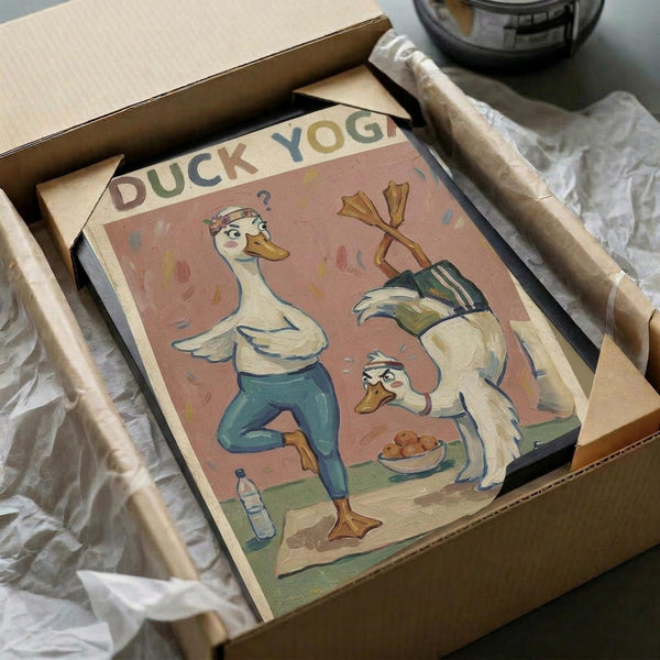 May include: Vintage-style poster artwork featuring two cartoon ducks doing yoga poses, titled "Duck Yoga" in colorful letters. One duck wears teal leggings while stretching, the other duck sits nearby with a bowl of snacks. The illustration has warm peachy-pink background tones with retro styling. The framed print sits in an open cardboard shipping box with white tissue paper padding, surrounded by wooden dowels on a textured surface.