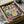 May include: Colorful folk art kitchen print in black frame, packed in cardboard shipping box with protective tissue paper. Artwork shows whimsical illustrated kitchen scene with potted plants on shelf, hanging utensils, decorative teapots and mugs in vibrant blues, yellows, pinks and greens. Print features hand-drawn style with floral patterns and botanical elements throughout kitchen items.