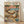 May include: Framed poster print leaning against white wall on light wood floor. Artwork shows two orange tabby cats relaxing on tropical beach - one in hammock wearing sunglasses and straw hat, another sitting beside wearing floral Hawaiian shirt and sunglasses. Scene includes palm trees, blue ocean, pink sunset sky, side table with fish plate and wine glass, scattered books and takeout containers on sand. Colorful letters spell "CAT NAP" at top. Natural wood frame surrounds vintage-style illus