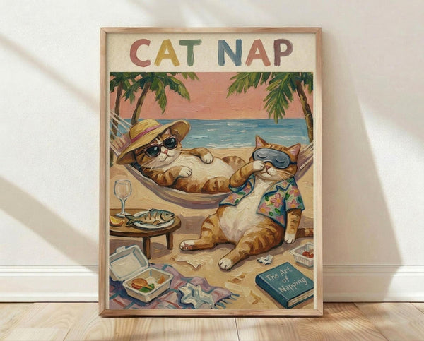 May include: Framed poster print leaning against white wall on light wood floor. Artwork shows two orange tabby cats relaxing on tropical beach - one in hammock wearing sunglasses and straw hat, another sitting beside wearing floral Hawaiian shirt and sunglasses. Scene includes palm trees, blue ocean, pink sunset sky, side table with fish plate and wine glass, scattered books and takeout containers on sand. Colorful letters spell "CAT NAP" at top. Natural wood frame surrounds vintage-style illus
