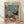 May include: Framed poster print leaning against white wall on light wood floor. Artwork shows cartoon cat DJ wearing sunglasses and floral shirt at turntables, golden retriever bartender in black tuxedo making cocktails. Colorful confetti and balloons fill blue background. Party spread includes cheese board, appetizers, disco balls, and cocktail glasses on pink surface below.