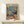 May include: Framed colorful poster showing a cat DJ wearing sunglasses and Hawaiian shirt at turntables, with a golden retriever bartender in tuxedo holding cocktail shaker. Scene includes party balloons, confetti, disco balls, cheese platter, and cocktails on table. Small white gift box with greenery sits on light wood shelf below the artwork against white wall.