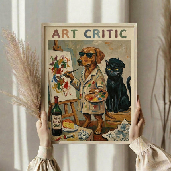 May include: Person holding framed poster showing brown dog wearing sunglasses and paint-splattered shirt, holding paintbrush and palette while standing next to easel with colorful abstract painting. Black cat with beret sits nearby. Scene includes wine bottle labeled "Vin Rouge Chat", cheese, and art supplies scattered around. Colorful text reads "ART CRITIC" at top. Light wooden frame against neutral background with dried pampas grass visible.