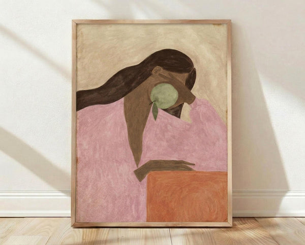 May include: Framed poster print featuring an abstract portrait of a woman with long dark brown hair wearing a pink top, holding a green apple. The artwork uses warm earth tones against a cream background. The print is displayed in a light wood frame, leaning against a white wall on light wooden flooring with soft natural lighting creating gentle shadows.