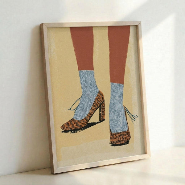 May include: Framed artwork showing two legs wearing rust-colored pants, light blue textured socks, and orange-brown patterned high-heeled mary jane shoes with ankle straps against a cream background. The illustration has a vintage mid-century style with bold colors and simple forms. The piece is displayed in a natural wood frame, leaning against a white wall.