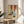 May include: Framed artwork showing legs wearing blue textured socks and leopard print heeled shoes with black straps against a yellow background, displayed on a round wooden side table next to a beige upholstered chair with white throw pillow, green plant visible in corner, natural sunlight streaming across white walls