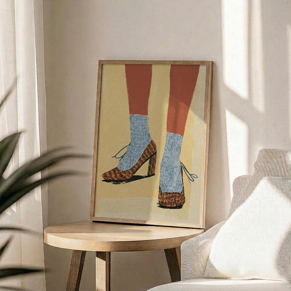 May include: Framed artwork showing legs wearing blue textured socks and leopard print heeled shoes with black straps against a yellow background, displayed on a round wooden side table next to a beige upholstered chair with white throw pillow, green plant visible in corner, natural sunlight streaming across white walls