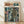 May include: Framed poster print leaning against white wall on light wood floor. Shows anime-style illustration of pilot in brown flight suit standing beside large green and tan military mecha robot in futuristic city street with neon signs. Japanese text at top in orange and teal colors with "Steel Lone Wolf" subtitle. Natural wood frame, soft lighting with shadows cast on textured white wall.