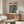 May include: A framed poster displaying a large military mech robot and pilot figure on a futuristic city street with neon signs and Japanese text. The artwork features detailed illustrations in muted greens and browns. The poster hangs above a mid-century modern walnut credenza with brass hardware in a minimalist living room setting with white walls, a curved white chair, glass coffee table, and warm natural lighting.