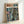 May include: Framed poster print leaning against white wall showing anime-style mecha robot pilot in brown armor standing in neon-lit cyberpunk city street. Artwork features detailed mechanical suit, glowing signs, wet pavement reflections in blues, greens, and pink tones. Light wooden frame on light surface.