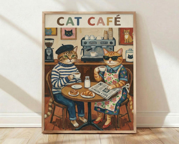May include: Framed poster print leaning against white wall on light wood floor. Artwork shows two anthropomorphic cats sitting at round cafe table - one tabby in black beret and striped shirt, another orange cat in sunglasses and floral dress. Behind them, coffee machine and cat portraits on wall. Colorful illustrated style with warm browns, blues, and oranges. Natural wood frame.