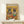 May include: Framed poster print displayed on light wood console against white wall, featuring two French bulldogs in colorful Mexican attire sitting at table with tacos, guacamole, margaritas and tequila bottles on vibrant yellow and orange background. Small white wrapped gift sits on shelf below frame.