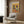 May include: Framed poster showing two French bulldogs in colorful Mexican attire sitting at a table with tacos, guacamole, and festive food on yellow and orange background, displayed above a cream upholstered chair with geometric pillow in modern room with wood paneling and gold floor lamp.