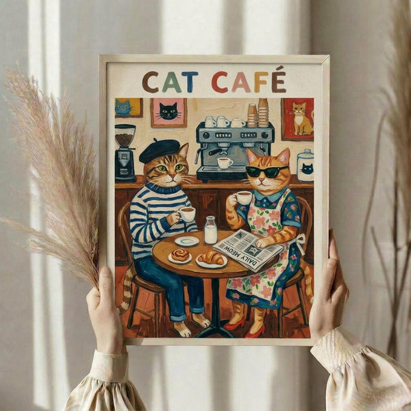 May include: Person holding framed poster showing two anthropomorphic cats sitting at cafe table with coffee cups and pastry. One cat wears striped shirt and beret, other wears floral apron and sunglasses. Background shows espresso machine, coffee grinder, and cat portraits on wall. Colorful text reads "CAT CAFÉ" at top. Light wooden frame, neutral wall backdrop, dried wheat stems visible on left side.