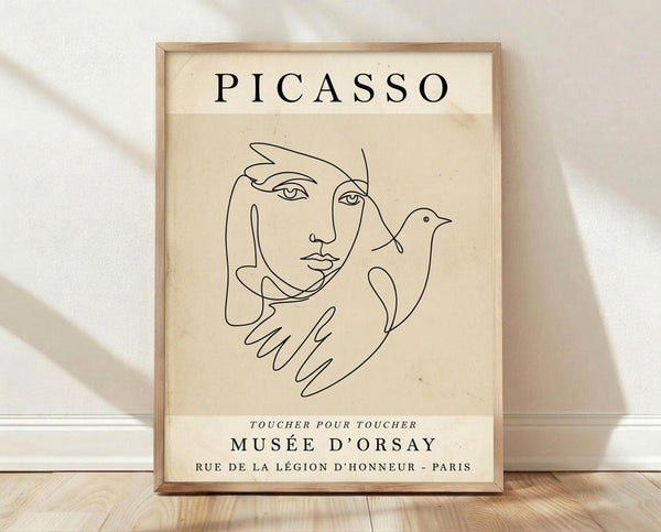 May include: Framed beige poster with black line drawing in Picasso's minimalist style showing a woman's face and hands holding a dove. Text reads "PICASSO" at top and "TOUCHER POUR TOUCHER MUSÉE D'ORSAY RUE DE LA LÉGION D'HONNEUR - PARIS" at bottom. Light wood frame leaning against white wall on wooden floor with soft shadows.