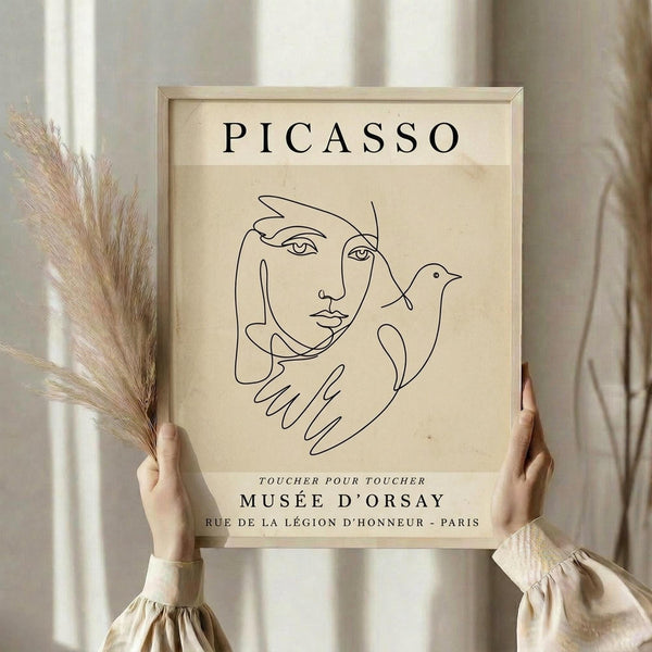 May include: Person in cream sweater holding framed beige poster featuring black line drawing of woman's face with hands holding dove, dried pampas grass visible to left, white curtained window background, natural wood frame